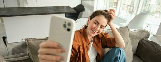 Porträt von Schön, glücklich jung weiblich Modell, Sitzung auf Couch mit Smartphone, nehmen Selfie auf Handy, Mobiltelefon Telefon, Herstellung heiter lächelnd Gesicht foto