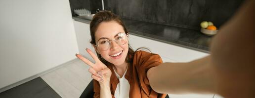 Selfie von stilvoll jung Frau, Lebensstil Blogger Erfassen ihr Wochenende beim heim, posieren zum Foto, erweitert Hand und hält Smartphone, lächelt und zeigt an vsign foto