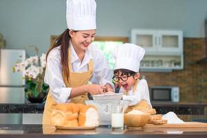 Süßer kleiner asiatischer Junge und schöne Mutter, die Teigmehl mit Siebsieb in der heimischen Küche auf dem Tisch sieben, um sich auf das Backen von Bäckerei und Kuchen vorzubereiten. thailändische kinder spielen mit mehl als koch lustig foto