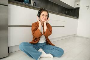 süß modern Mädchen auf Küche Fußboden beim heim, Hören Musik- im kabellos Kopfhörer, haben Spaß foto