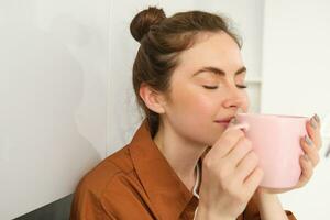 schließen oben Porträt von schön Brünette Frau mit Tasse von Kaffee, Trinken aromatisch Getränk von Becher, lächelnd von Vergnügen, Sitzung im Küche beim Zuhause foto