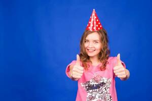 Feier und Party. haben Spaß. jung ziemlich Frau im Geburtstag Hut ist Lachen und zeigen Daumen hoch. bunt Studio Porträt mit Blau Hintergrund. foto