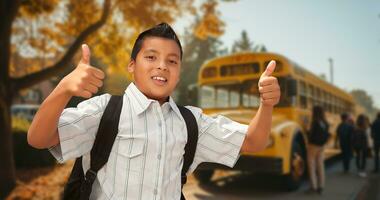 aufgeregt jung spanisch Junge tragen ein Rucksack geben zwei Daumen oben auf Campus in der Nähe von ein Schule Bus foto