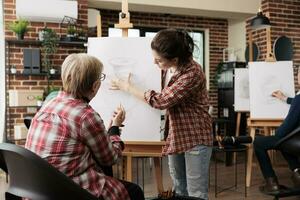 weiblich Kunst Lehrer Stehen in der Nähe von Staffelei erklären Senior Frau Wie zu zeichnen immer noch Leben Objekte mit Bleistift, Lehren Menschen Zeichnung Techniken während Gruppe Kunst Klasse. Lernen Neu Dinge im Pensionierung foto