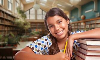 glücklich jung spanisch Mädchen mit ein Bleistift und ein Stapel von Bücher tun Hausaufgaben im ein Bibliothek foto