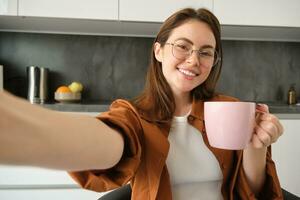 Porträt von schön jung Frau im Gläser, nehmen Selfie im Küche mit Tasse von Morgen Kaffee, Herstellung Fotos zum ihr Täglich Routine Lebensstil Blog