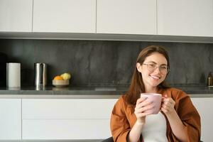 Porträt von glücklich jung Frau im Gläser, Sitzung beim Zuhause im Küche, halten Tasse von Kaffee, Trinken Lieblings Tee, entspannend drinnen auf kalt Herbst Tag foto