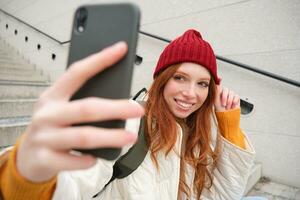 Handy, Mobiltelefon Telefon und Menschen Lebensstil. stilvoll Rothaarige Mädchen nimmt Selfie auf ihr Smartphone, posiert zum Foto mit Handy, Mobiltelefon Telefon im Hand, lächelt glücklich, sitzt auf Treppe draußen