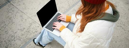 Rückseite Aussicht von Mädchen Schüler Tippen auf Laptop Klaviatur, leer schwarz Bildschirm zum Webseite Anwendung Werbung, Sitzung auf Treppe draußen, verbindet zu Öffentlichkeit W-lan foto