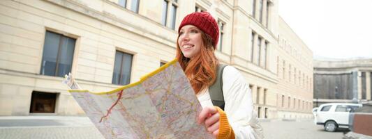 schön Rothaarige Frau, Tourist mit Stadt Karte, erforscht Besichtigung historisch Wahrzeichen, Gehen um alt Stadt, lächelnd glücklich foto