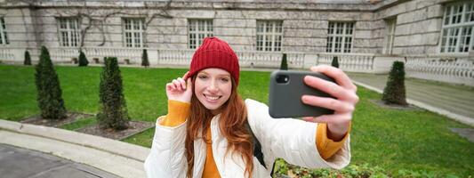 schön lächelnd Ingwer Mädchen im Hut, posieren zum Foto auf Handy, Mobiltelefon Telefon, nimmt Selfie im Park mit süß Gesicht Ausdruck