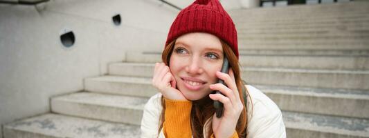 schön lächelnd Rothaarige weiblich Modell, sitzt auf Straße und Gespräche auf Handy, Mobiltelefon Telefon, Verwendet Smartphone App zu Anruf im Ausland, Lachen während Telefon Konversation foto