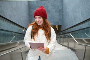 schön Rothaarige weiblich Modell- posieren im Stadt, Gehen oben Treppe mit Digital Tablette, mit Gadget zu planen ihr Route, lesen während gehen irgendwo foto