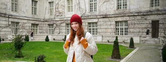 Porträt von lächelnd Mädchen, Backpacker im Mantel und rot Hut, tragen warm Kleider zum Tourist Ausflug um Europa im Winter, Gehen um historisch Gebäude foto