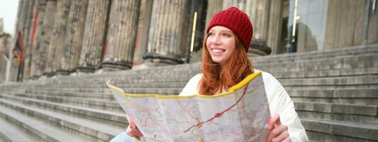 Porträt von jung Rothaarige Frau, Tourist sitzt mit Papier Karte und sieht aus zum ein Route zu Tourismus Attraktion, ruht auf Treppe draußen foto