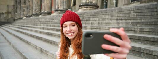 jung Rothaarige Tourist nimmt Selfie im Vorderseite von Museum auf Stufen, hält Smartphone und sieht aus beim Handy, Mobiltelefon Kamera, macht Foto von Sie selber mit Telefon