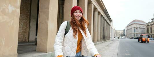 lächelnd Rothaarige europäisch Mädchen fährt Öffentlichkeit Escooter, Tourist erforscht Stadt, Fahrten im Stadt Center foto