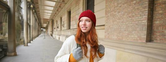 Rucksack Tourist. glücklich Rothaarige Mädchen Spaziergänge im Stadt mit ein Tasche, geht auf Reise, erforscht Stadt, trägt rot Hut und Handschuhe foto