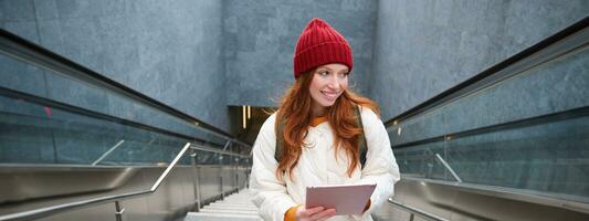 schön Rothaarige weiblich Modell- posieren im Stadt, Gehen oben Treppe mit Digital Tablette, mit Gadget zu planen ihr Route, lesen während gehen irgendwo foto