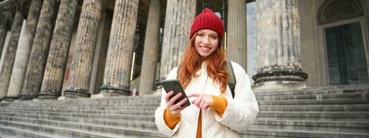 Porträt von jung Rothaarige Frau halten Handy, Mobiltelefon Telefon, mit Smartphone Anwendung, Gehen im Stadt Center, mit Karte Anwendung oder SMS ein Botschaft foto