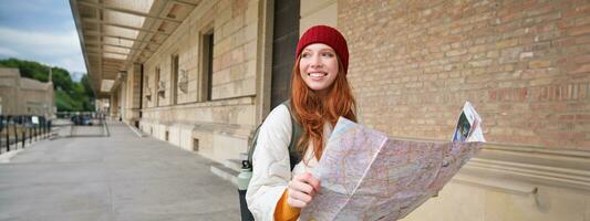 abenteuerlich Rothaarige Mädchen Spaziergänge im Stadt, Dorf mit Papier Karte, erforscht Stadt wie Tourist, sieht aus zum Beliebt Tourismus Sehenswürdigkeiten, sieht aus um aufgeregt und lächelt foto