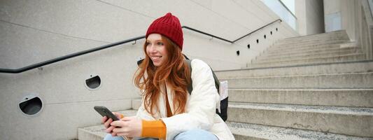 stilvoll europäisch Mädchen mit rot Haar, sitzt auf Öffentlichkeit Treppe mit Smartphone, setzt online Befehl, sendet Botschaft auf Handy, Mobiltelefon Telefon Sozial Anwendung, lächelt glücklich foto