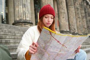 Tourismus und Lebensstil Konzept. jung Rothaarige Frau suchen beim Stadt Karte, Pläne ein Route zum Besichtigung Tag, sitzt draußen auf Treppe und ruht foto