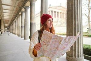 lächelnd jung Rothaarige Frau im rot Hut, sieht aus beim Papier Karte zu aussehen zum Tourist Attraktion. Tourismus und Menschen Konzept. Mädchen erforscht Stadt, versucht zu finden Weg foto