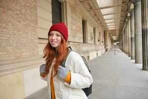 lächelnd Rothaarige Mädchen mit Rucksack, Spaziergänge im Stadt und tut Besichtigung, erkunden Beliebt Sehenswürdigkeiten auf ihr Tourist Reise um Europa foto