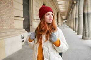 Rucksack Tourist. glücklich Rothaarige Mädchen Spaziergänge im Stadt mit ein Tasche, geht auf Reise, erforscht Stadt, trägt rot Hut und Handschuhe foto