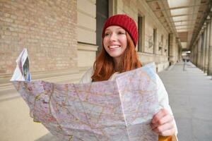 lächelnd jung Rothaarige Frau im rot Hut, sieht aus beim Papier Karte zu aussehen zum Tourist Attraktion. Tourismus und Menschen Konzept. Mädchen erforscht Stadt, versucht zu finden Weg foto