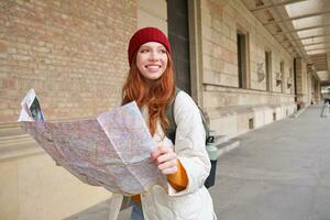 abenteuerlich Rothaarige Mädchen Spaziergänge im Stadt, Dorf mit Papier Karte, erforscht Stadt wie Tourist, sieht aus zum Beliebt Tourismus Sehenswürdigkeiten, sieht aus um aufgeregt und lächelt foto