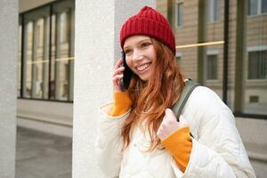 Porträt von lächelnd Ingwer Mädchen mit rot Hut, steht auf Straße mit Rucksack, Ringe jemand auf Telefon Anwendung, Gespräche auf Handy, Mobiltelefon, Verwendet Smartphone foto