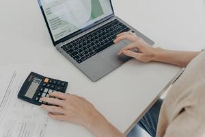 Unerkennbare Frau sitzt am Schreibtisch mit Laptop-Rechner-Papieren berechnet monatliche Ausgaben foto