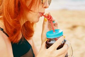 Nahansicht von rot Haar Frau Lippen Trinken alkoholisch Cocktail mit ein Stroh. Meer Strand im das Hintergrund. Dame im schwarz Badeanzug auf Ferien im das Tropen. Foto Karte mit Kopieren Raum.