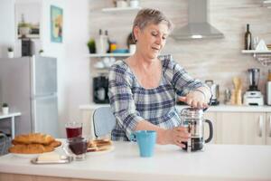 Senior Frau genießen ein Becher von Kaffee von Französisch Drücken Sie während Frühstück. Alten Person im das Morgen genießen frisch braun Cafe Espresso Tasse Koffein von Jahrgang Becher, Filter entspannen Erfrischung foto