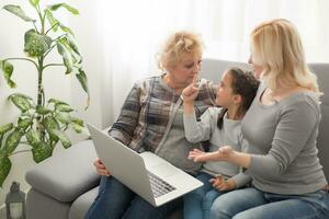 glücklich drei Generationen von Frauen sitzen entspannen auf Couch Lachen Aufpassen komisch Video auf Laptop, lächelnd positiv Frauen Oma, Mutter und Tochter haben Spaß sich ausruhen auf Sofa genießen Film auf Computer foto