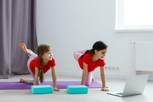 Zwei kleine Mädchen praktizieren Yoga, Stretching, Fitness per Video auf dem Notebook. entferntes Online-Bildungstraining, Aerobic zu Hause. gesunder lebensstil, coronavirus, bleib zu hause. Kindersport Quarantäne zu Hause. foto
