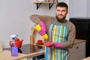 jung gut aussehend bärtig Mann im das Küche, tragen Schürze und Rosa Handschuhe reinigt das Kochen Oberfläche mit Waschmittel foto