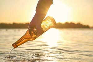 Freiwillige Hand im lila Handschuh nimmt aus ein benutzt Glas Flasche von das Fluss. Sonnenuntergang, Grün Bäume. Menschen und Ökologie. Flussufer Verschmutzung. Nahansicht. foto