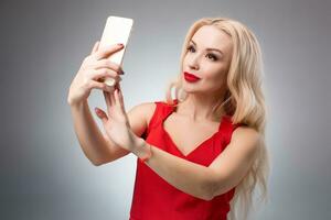 Porträt von ein schön erfolgreich lächelnd Mädchen tun Selfie im rot Kleid auf Licht Hintergrund foto
