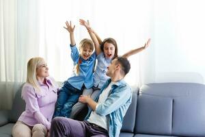 heiter jung Familie mit Kinder Lachen Sitzung auf Couch zusammen, Eltern mit Kinder genießen unterhaltsam beim Zuhause foto