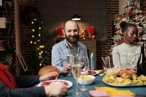 jung Mann genießen festlich Weihnachten Abendessen mit vielfältig Menschen, Treffen mit Familie um das Tabelle zum Weihnachten Vorabend. vielfältig Personen haben Spaß während Winter Jahreszeit Feier, gemütlich Dekor. foto