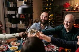 vielfältig Menschen Klirren Wein Brille beim Weihnachten Vorabend Abendessen, feiern Urlaub Veranstaltung mit Alkohol und hausgemacht Lebensmittel. freunde und Familie Sprichwort Prost mit Getränke beim Tisch, gemütlich Dekorationen. foto