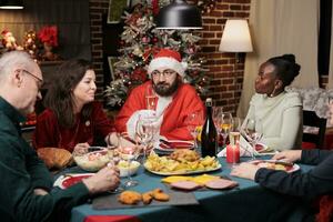 glücklich Santa claus Mann Herstellung Menschen Lachen beim Abendessen Tisch, feiern Weihnachten Urlaub zusammen beim heim. Senior Menschen, freunde und Familie Versammlung in der Nähe von Santa zu sagen Geschichten und haben Spaß. foto