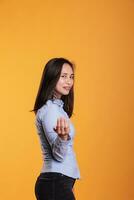 attraktiv asiatisch Frau einladend Sie zu Kommen Sie Hier mit ein freundlich Lächeln auf ihr zuversichtlich Ausdruck, posieren im Studio Über Gelb Hintergrund. froh jung Erwachsene ist Berufung aus mit ein Finger Geste foto