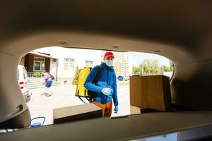 Essen Lieferung Mann im ein schützend Maske und Handschuhe mit ein thermo Rucksack in der Nähe von ein Auto während das Quarantäne Zeitraum foto