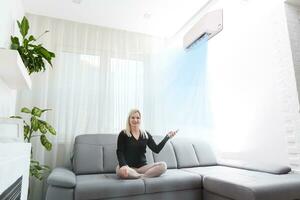 jung Frau Schalten auf Luft Conditioner während Sitzung auf Sofa in der Nähe von Weiß Mauer foto