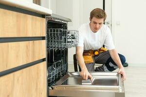 Techniker oder Arbeiter im Uniform installiert Geschirrspüler in das Küche Möbel. Handwerker tragen Arbeiter passen reparieren Instandhaltung von Geschirrspüler. Meister im schützend Handschuhe Fix Geschirrspüler. foto