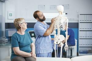 Krankenschwester und im Ruhestand geduldig suchen beim Wirbelsäule Kabel auf Mensch Skelett zum osteopatisch Untersuchung. medizinisch Assistent zeigen beim Rücken Knochen zu erklären Diagnose zu Senior Frau zum Gesundheitspflege. foto